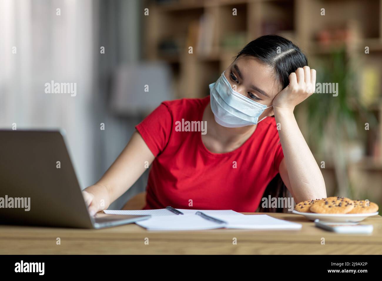 Unglückliche junge koreanische Dame in Schutzmaske sitzt am Tisch mit Laptop in der Zimmereinrichtung Stockfoto