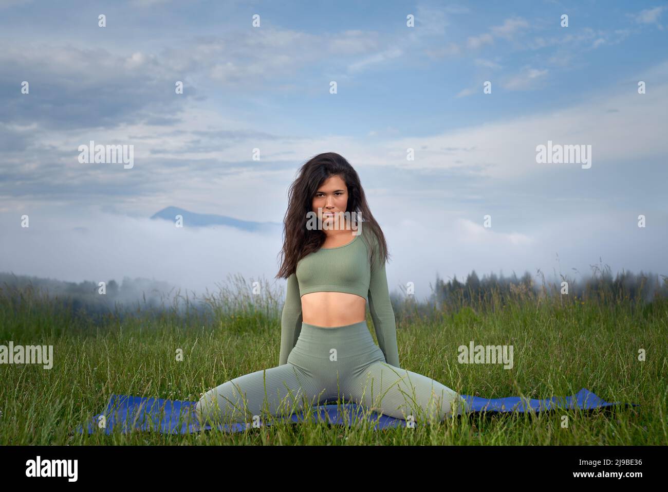 Schöne junge Frau mit dunklem Haar, die sich auf einer Yogamatte mit Bergen im Hintergrund ausdehnt. Aktive weibliche Person, die in der Freizeit Aktivitäten im Freien genießt. Stockfoto