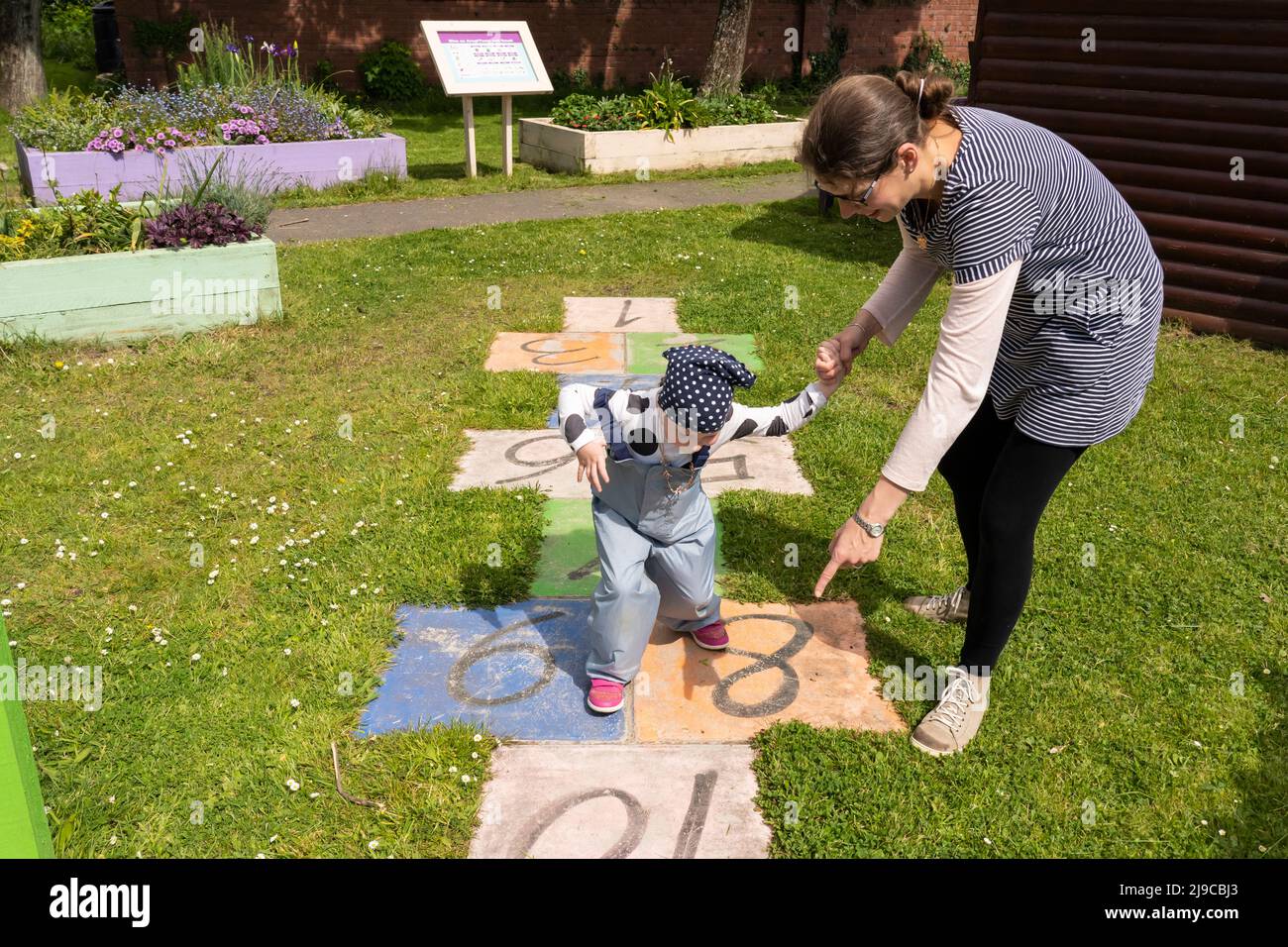 Eine Mutter lehrt ihre 4-jährige Tochter im Mai in Kidwelly Sensory Garden, Kidwelly, Carmarthen, Wales, wie man hopscotch spielt Stockfoto
