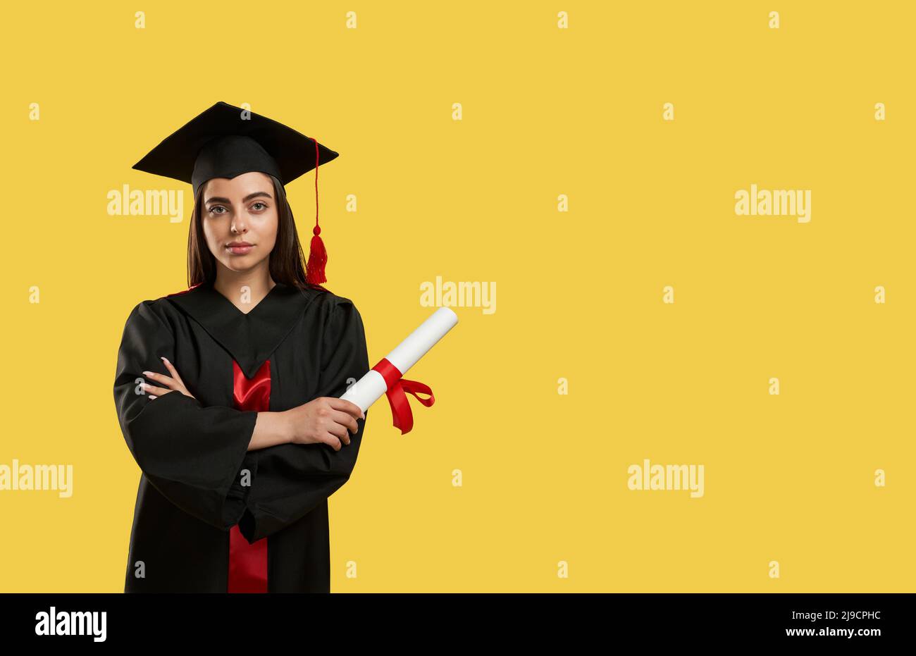 Vorderansicht des Mädchens, das mit gekreuzten Händen steht und Diplom hält. Brunette, die ihr Studium abschloss, mit Leichenbrett und Abschlusskleid, mit Blick auf die Kamera. Konzept der Jugend. Stockfoto