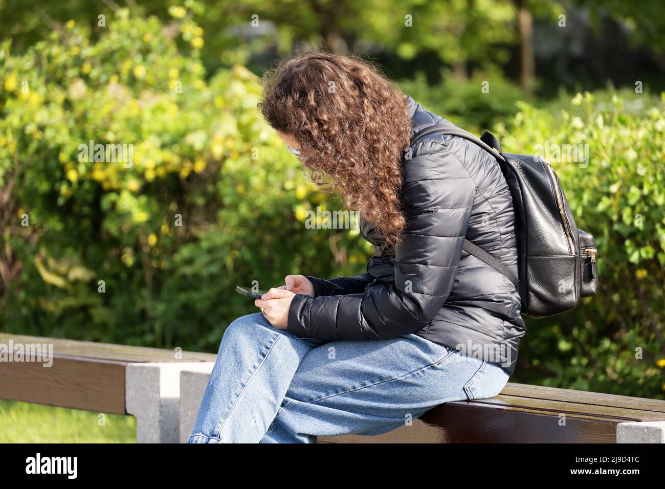 Mädchen mit lockigen Haaren trägt Jeans und wasserdichte Nylonjacke sitzt mit einem Smartphone auf der Bank im Frühling Park Stockfoto