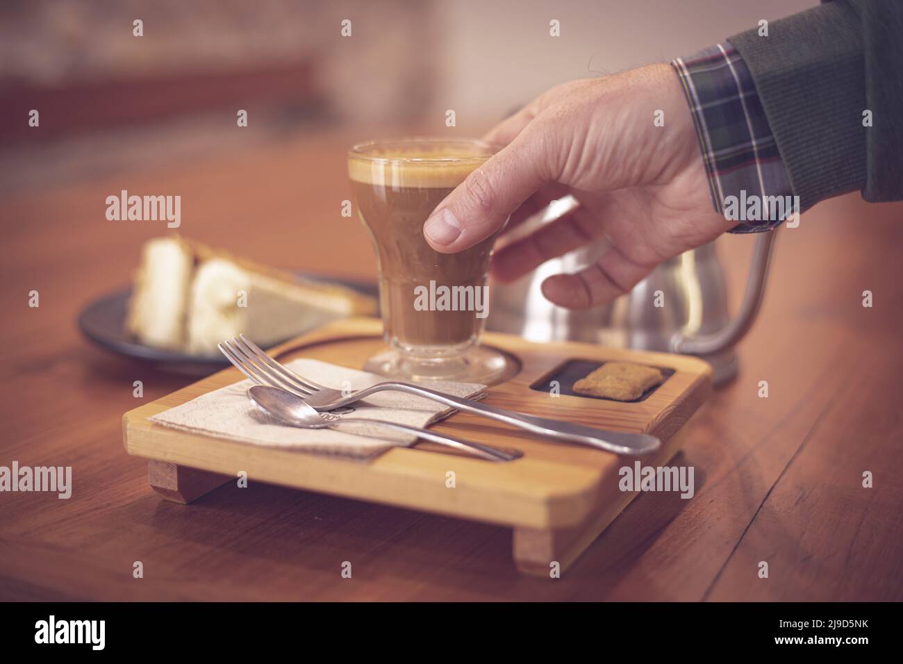 Stillleben im Zusammenhang mit Kaffee, wo ein Mann Hand nimmt eine Tasse Kaffee in einer Bar mit seinem Besteck und einem Karottenkuchen im Hintergrund. Cafe Latte. Stockfoto