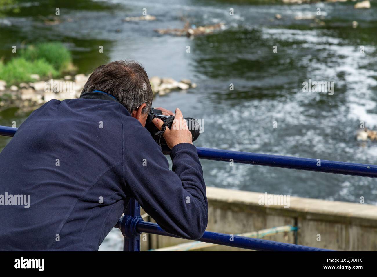Ein männlicher Fotograf lehnt sich über Geländer, um ein Bild vom Fluss Aire in Saltaire, Yorkshire, zu machen. Stockfoto