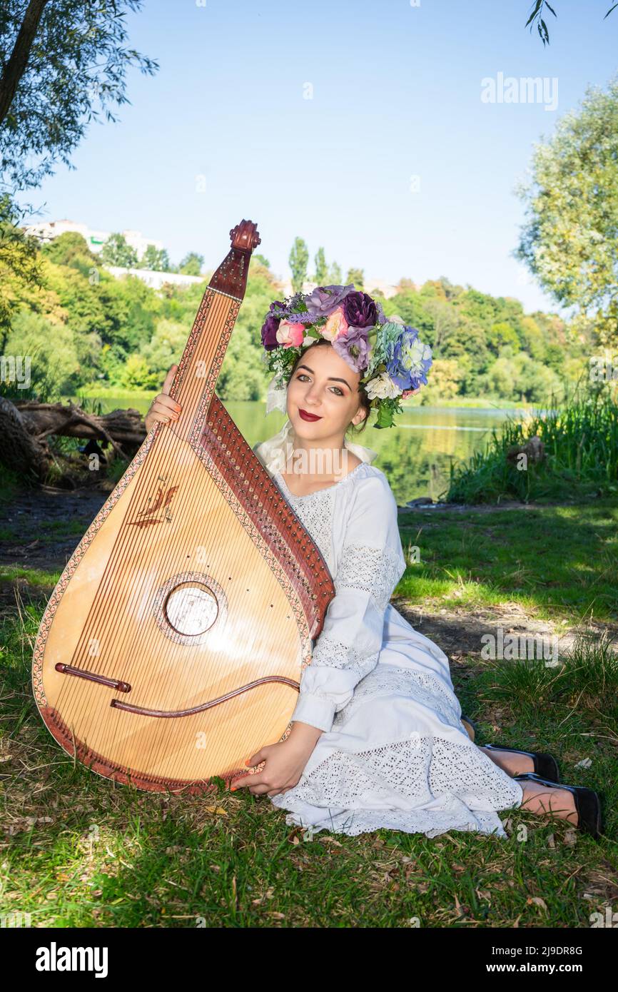 Junge ukrainische Frau, die in Nationaltracht am Fluss sitzt und ein ukrainisches Bandura-Instrument in der Hand hält. Stockfoto