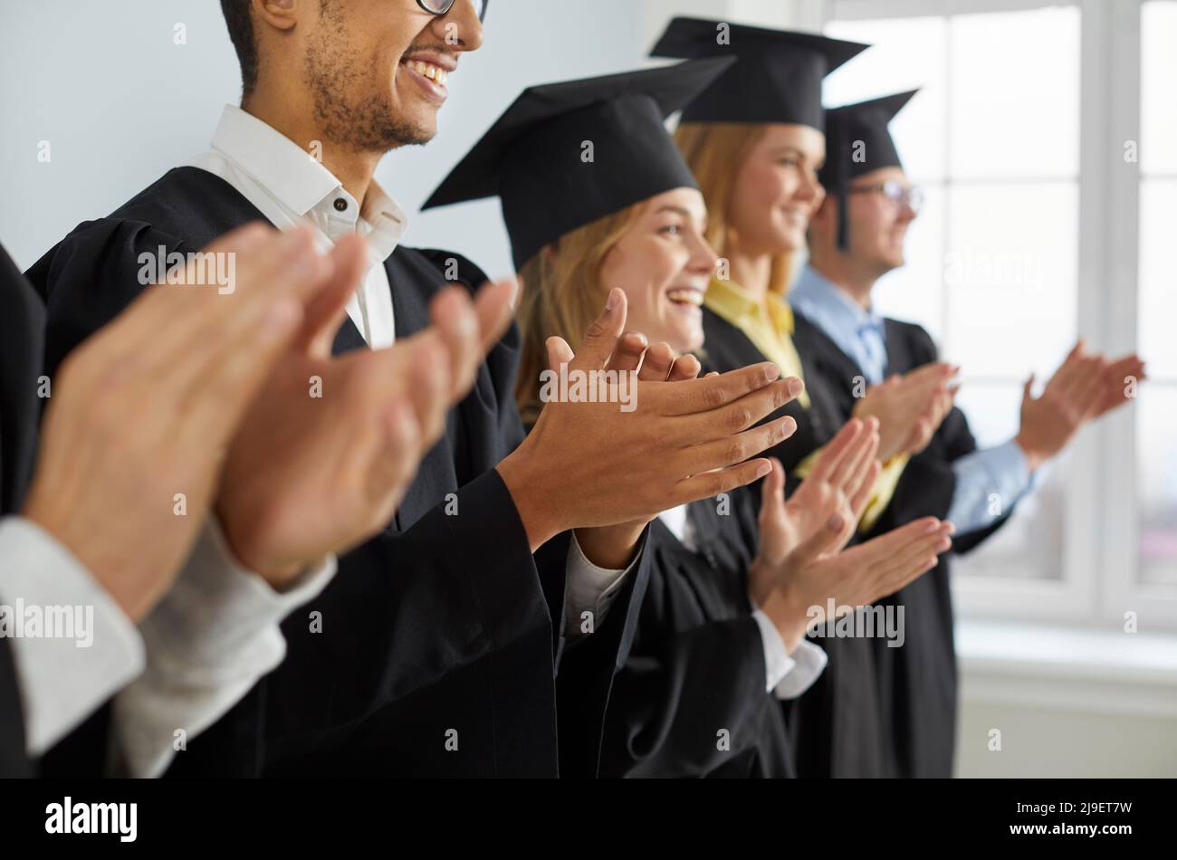Glückliche Absolventen in Kappen und Kleidern applaudieren bei der Abschlussfeier der Hochschule oder Universität Stockfoto