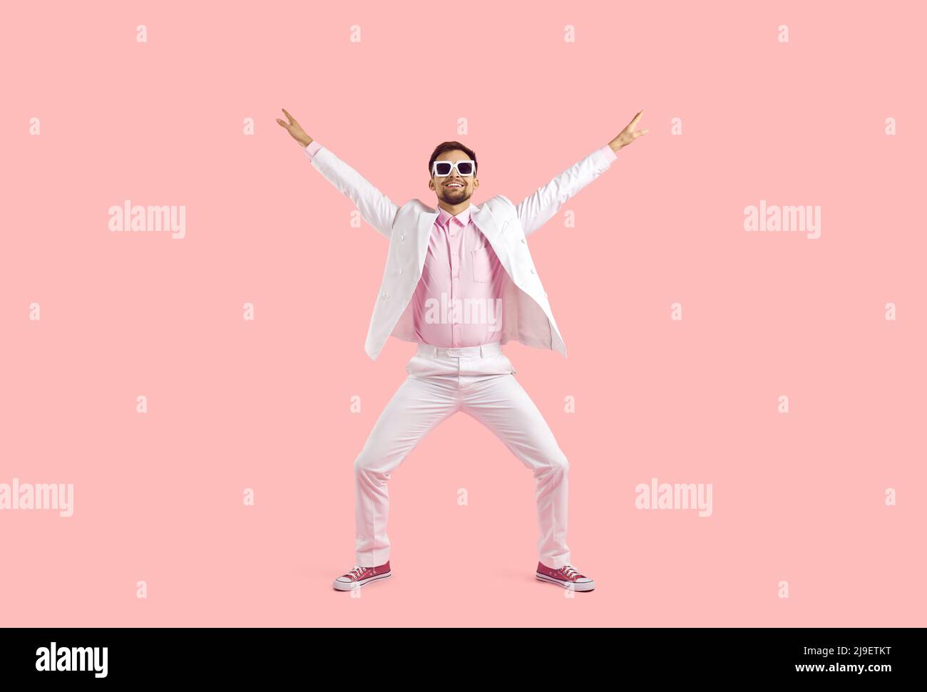 Foto in voller Länge von einer jungen Fashionista, die im Studio tanzt. Fröhlicher, hübscher junger Mann in einem weißen Anzug, einem rosa Hemd, pinken Turnschuhen und einer Sonnenbrille Stockfoto
