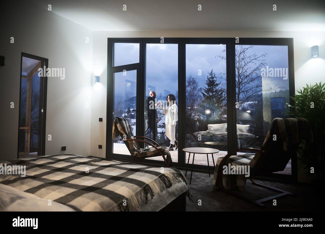 Schöne Aussicht auf Mann und Frau beim Toasten mit Wein hinter Panoramafenster des skandinavischen Hauskahnhauses. Innenraum des Holzhauses Zimmer mit Stühlen, Bett und verliebtes Paar außerhalb des Gebäudes. Stockfoto