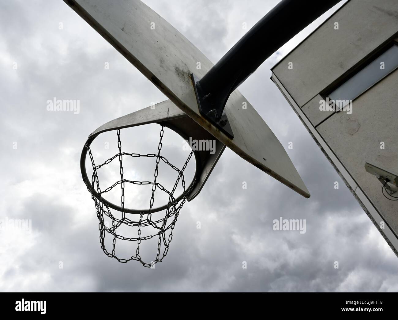 Ein vandalismussicherer Basketballkorb mit eisernen Ketten vor einem düsteren bewölkten Himmel, neben einem Betongebäude mit einer Überwachungskamera an der Wand Stockfoto