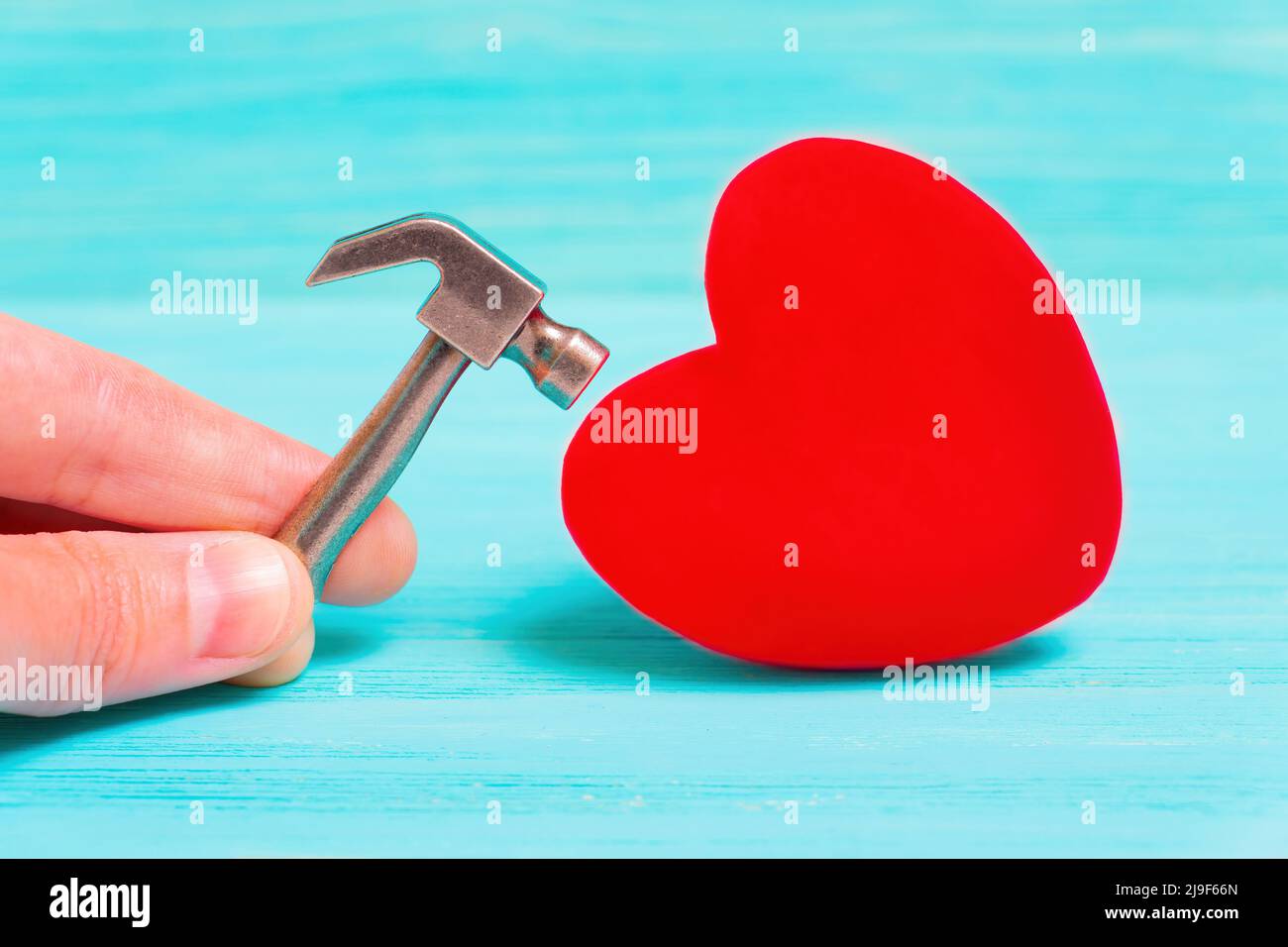 Treffen einer großen roten Herzform mit einem Miniatur-Stahlhammer isoliert auf einem türkisfarbenen Holzhintergrund. Herzfixierung. Stockfoto