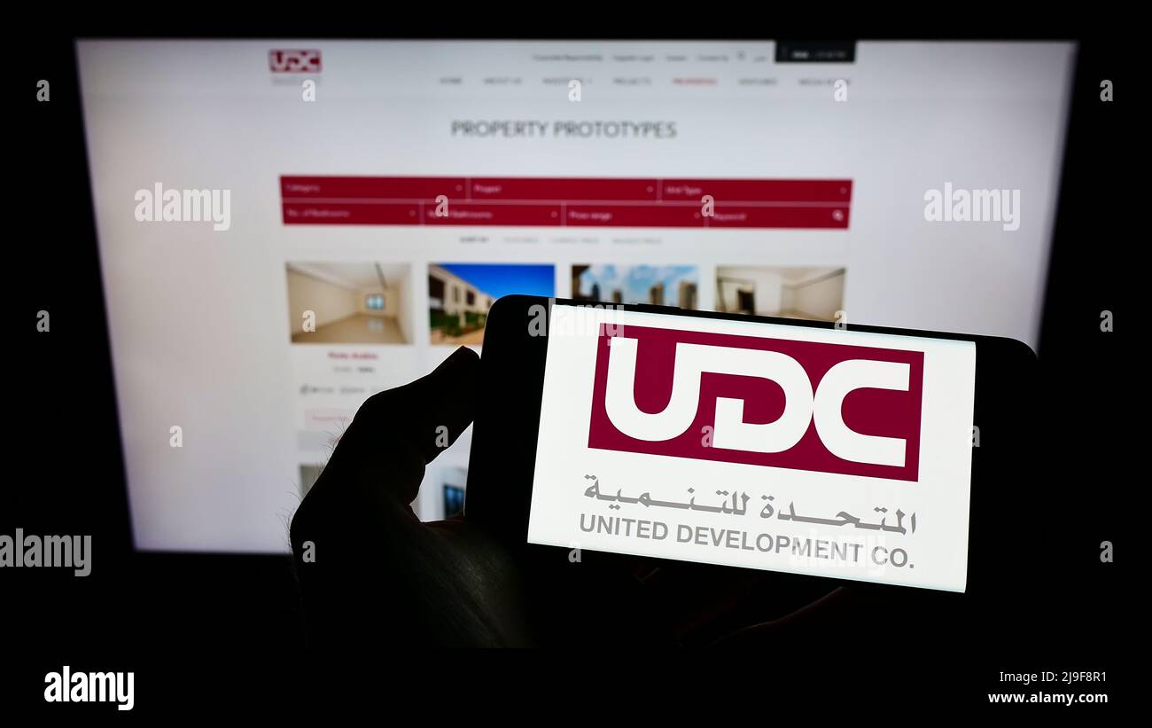 Person mit Mobiltelefon und Logo der United Development Company Q.P.S.C. (UDC) auf dem Bildschirm vor der Business-Webseite. Konzentrieren Sie sich auf die Telefonanzeige. Stockfoto