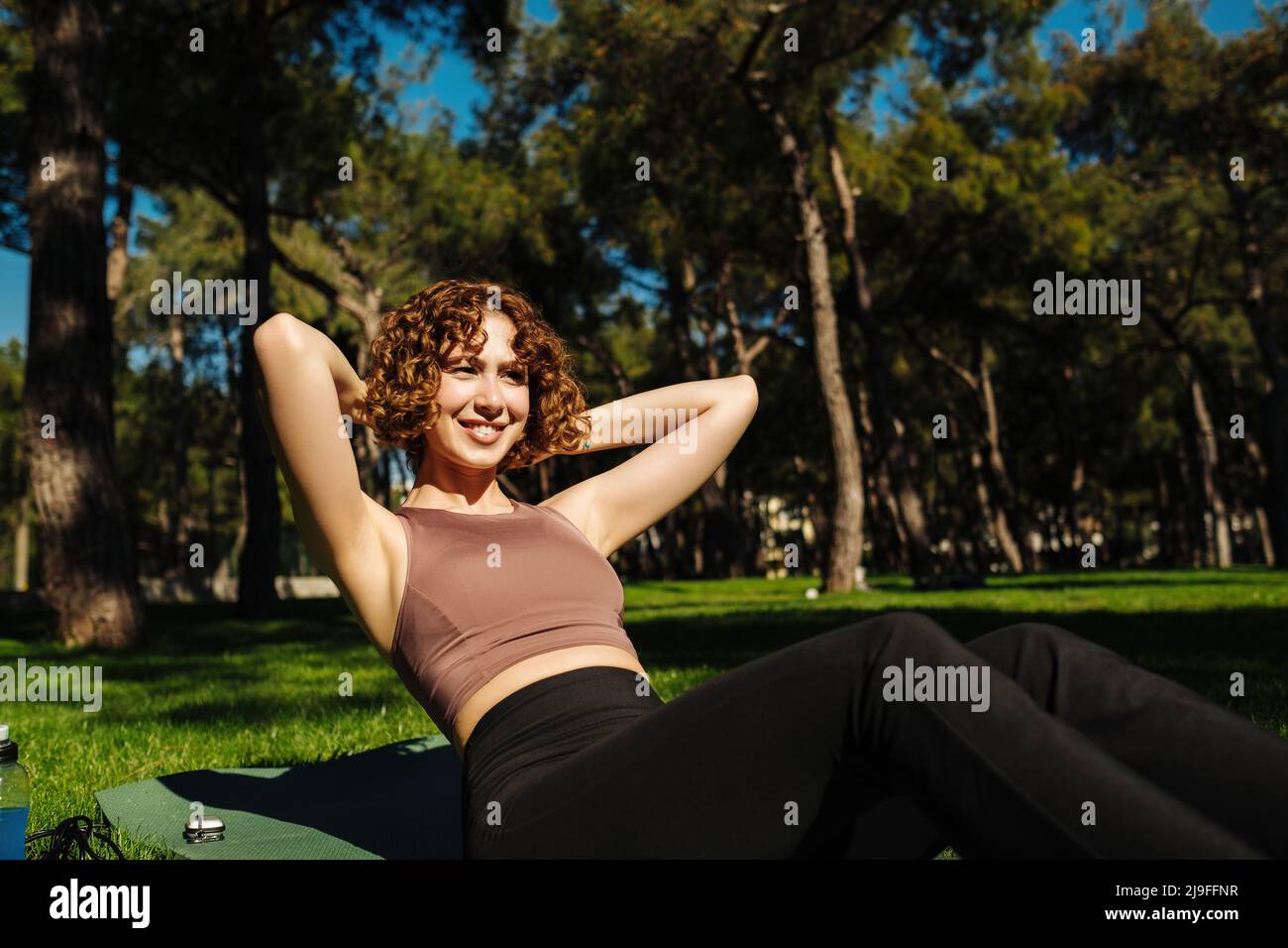 Sportlich lächelnde Rothaarige Frau trägt Sport-BH und macht Sit Ups abs Crunches trainieren im Freien auf Yoga-Matte liegen. Sport-Workout-Routine, morgens phys Stockfoto