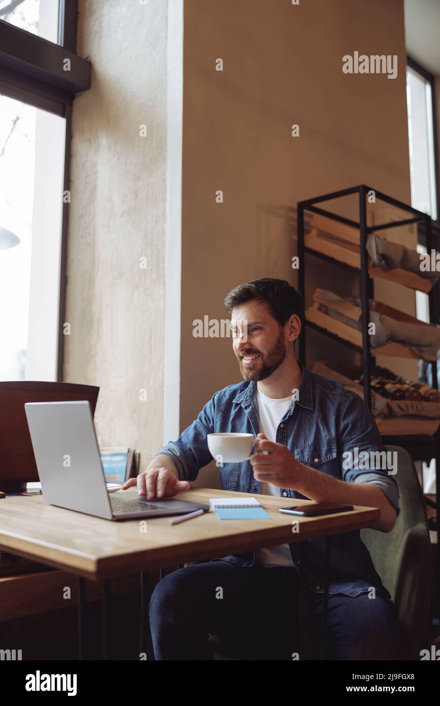 Fröhlicher kaukasischer junger attraktiver Mann mit einer Tasse Kaffee, der im Café am Laptop sitzt. Videoanruf. Stockfoto