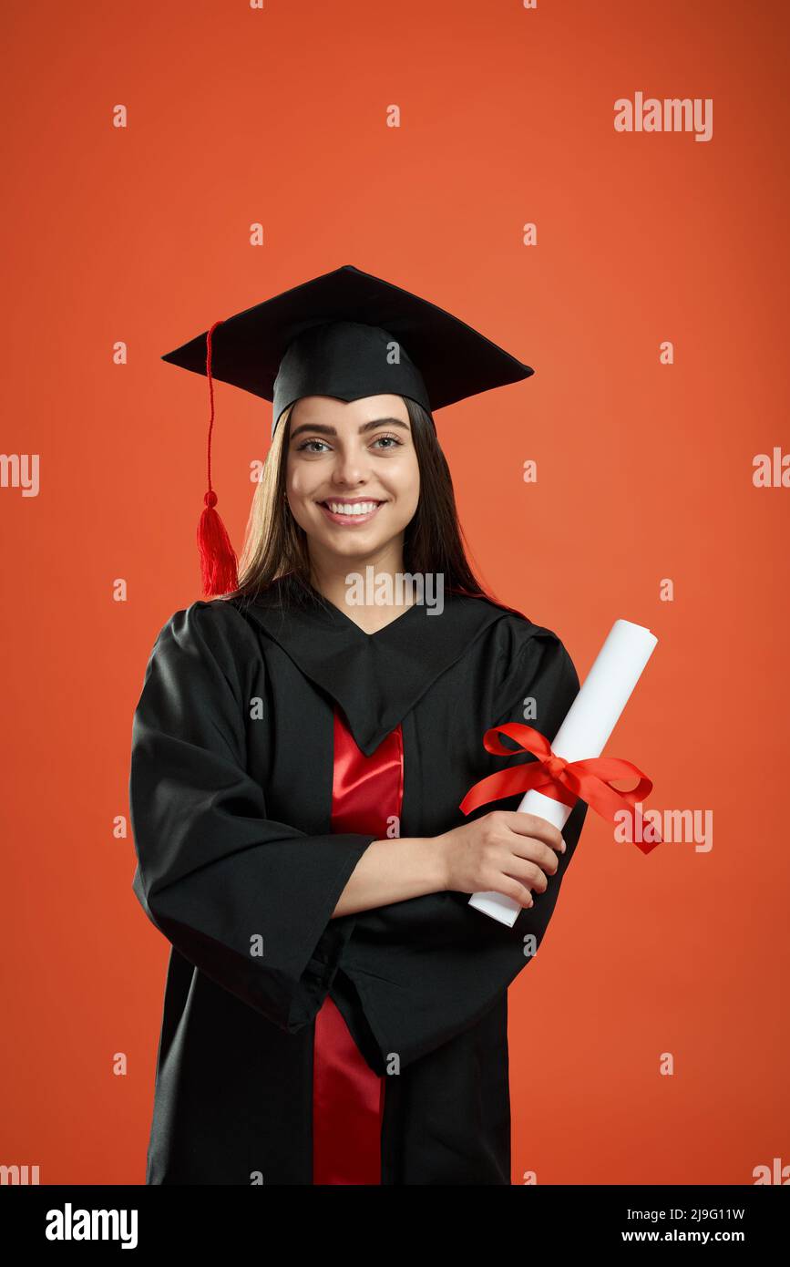 Vorderansicht eines hübschen Mädchens, das sein Studium an der Hochschule, Universität und Hochschule abschloss. Brunette junge weibliche stehend mit gekreuzten Händen, hält Diplom, lächelnd. Konzept der Jugend. Stockfoto