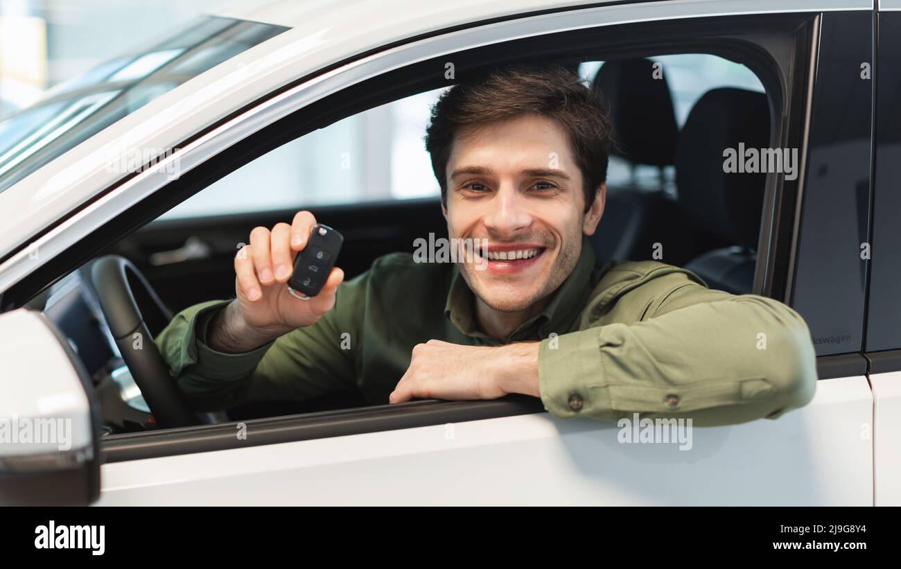 Glücklicher junger kaukasischer Mann, der Schlüssel aus dem Auto zeigt, im Autosalon sitzt und im Autohaus ein nagelneues Auto kauft Stockfoto