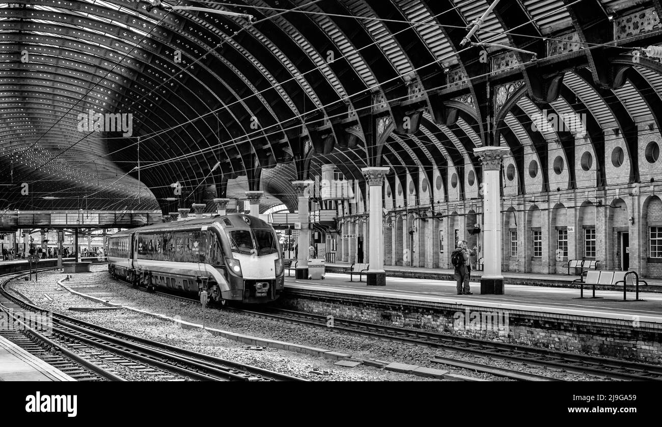 Ein moderner Zug steht an einer historischen Plattform mit Säulen und unter einem verzierten eisernen Baldachin. Passagiere sind auf dem Bahnsteig. Stockfoto