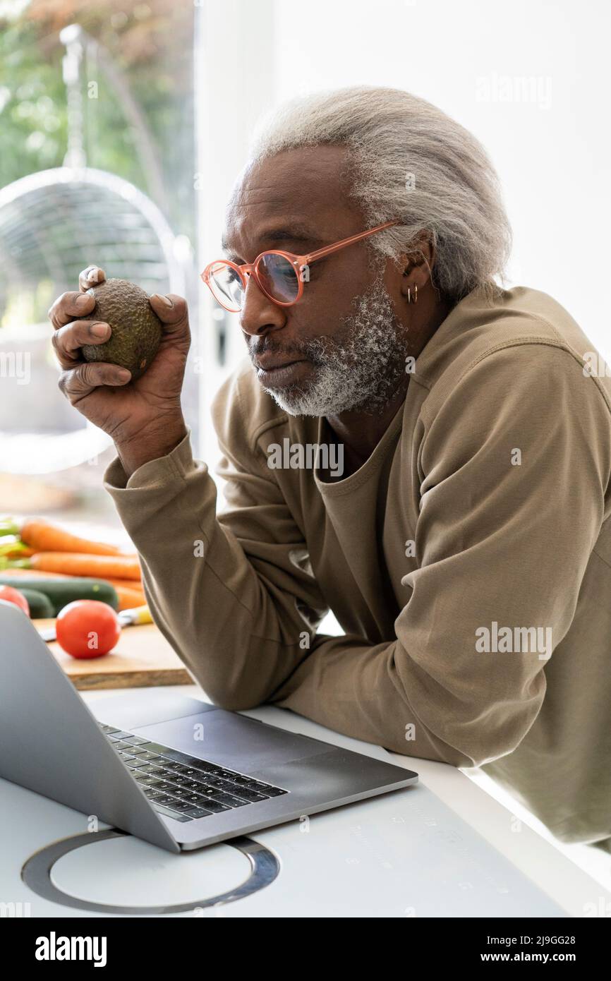 Ein älterer Mann schaut sich das Rezept auf dem Laptop an, während er sich auf die Küchenarbeitsfläche stützt Stockfoto