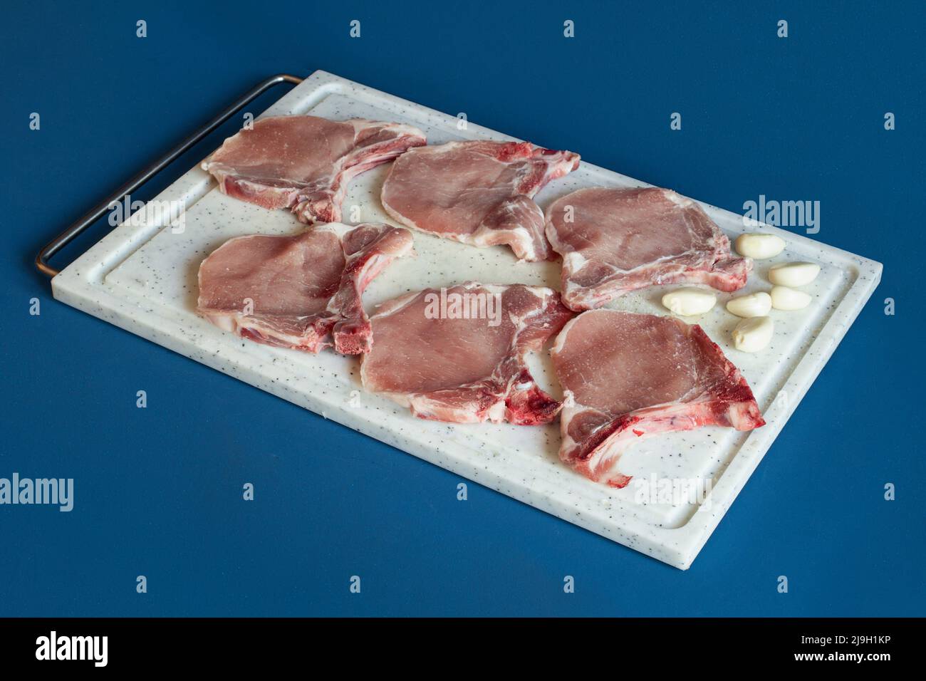 Schweinelende Koteletts mit einer Handvoll Knoblauchzehen auf einem Schneidebrett über einem farbenprächtigen blauen Studiohintergrund, bereit zum Kochen. Rohes Fleisch. Stockfoto