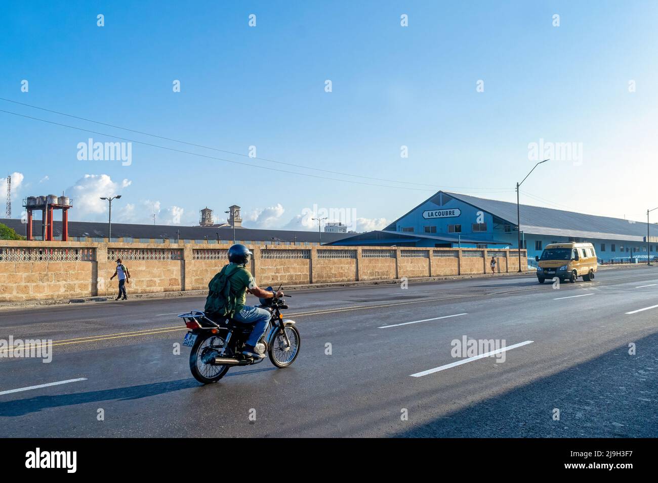 Ein Mann fährt an einem langen Zaun ein Motorrad auf einer Stadtstraße. Im Hintergrund befindet sich das Lagergebäude La Coubre. Stockfoto