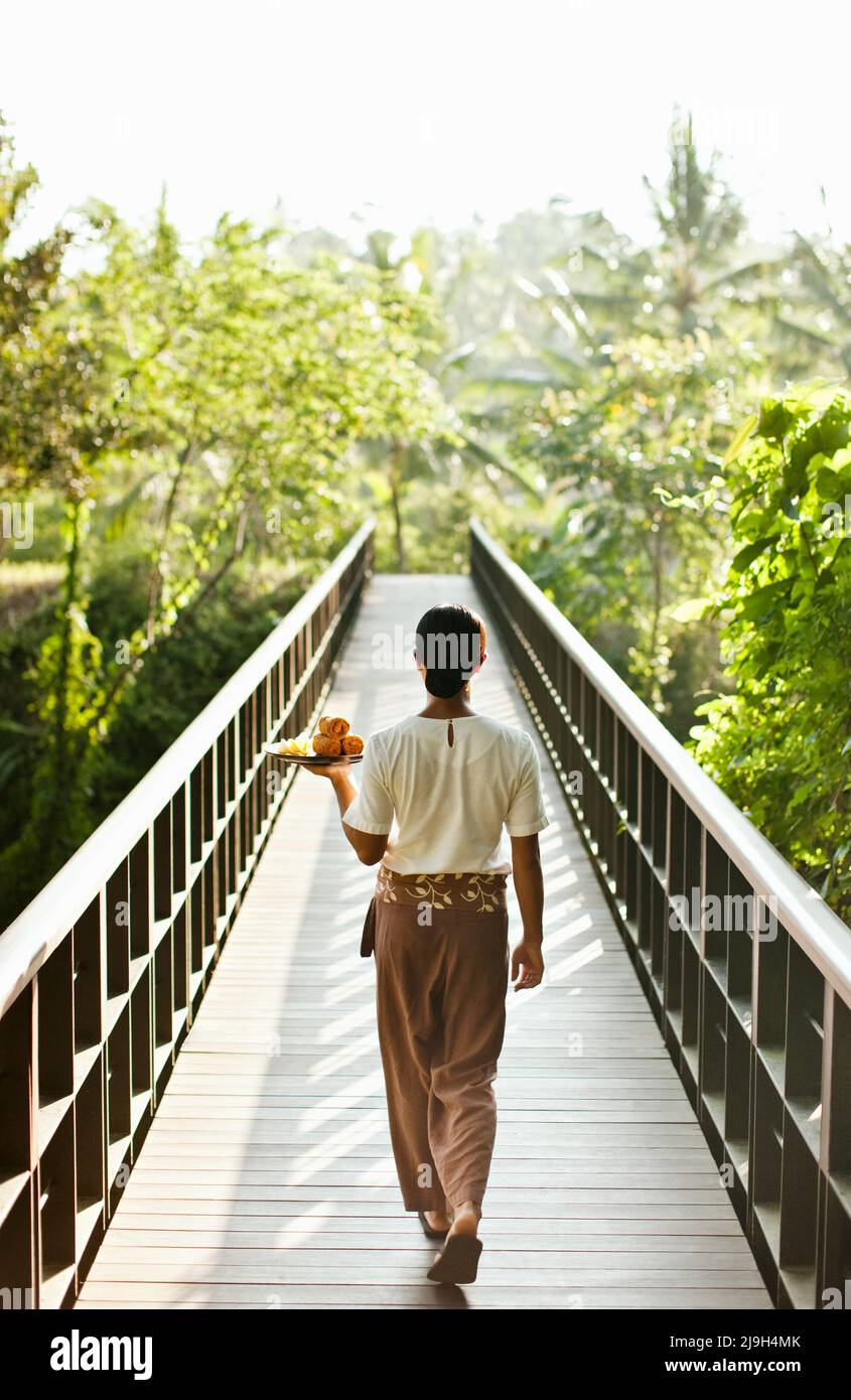Eine Frau trägt ein Tablett mit kalten Handtüchern über die Brücke, die das Resort mit dem Kayumanis Spa, Kayumanis Ubud, Bali, Indonesien verbindet. Stockfoto