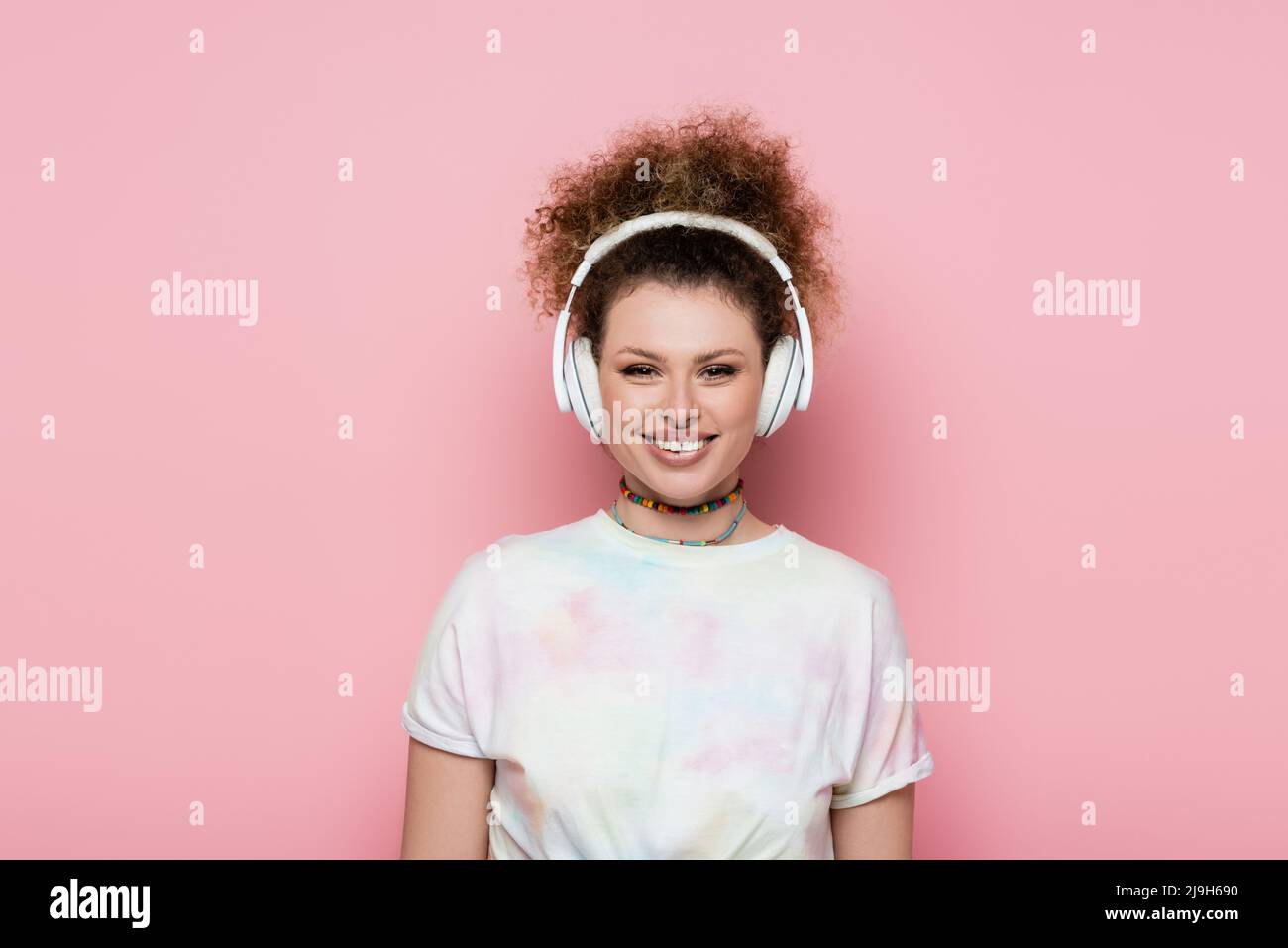 Lächelnde Frau in T-Shirt und Kopfhörer isoliert auf rosa Stockfoto