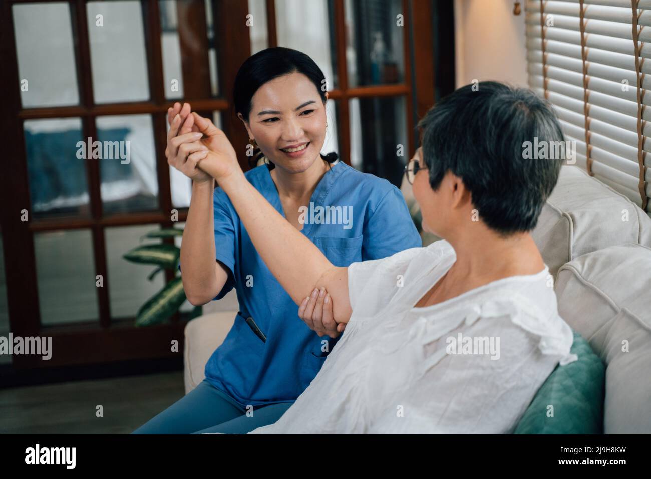 Entzückende weibliche Betreuerin in blauer Uniform lächelnd und streckend Arm einer älteren asiatischen Frau, während sie auf dem Sofa gegen das Fenster im Wohnzimmer zu Hause sitzt Stockfoto