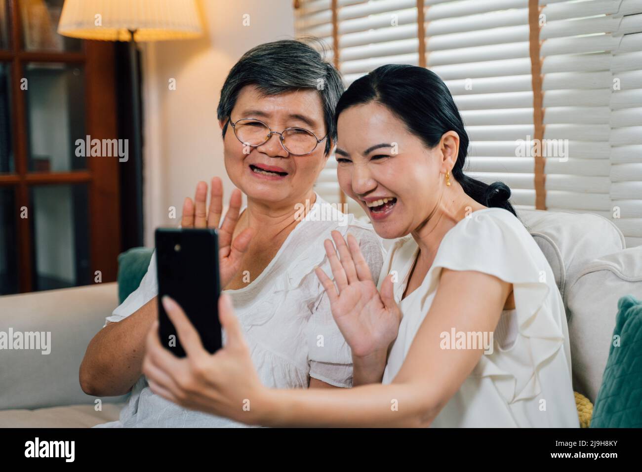 Fröhliche asiatische ältere Mutter und Erwachsene Tochter lächeln und winken die Hände, während sie tagsüber auf dem Sofa sitzen und Videoanruf über Mobiltelefon tätigen Stockfoto