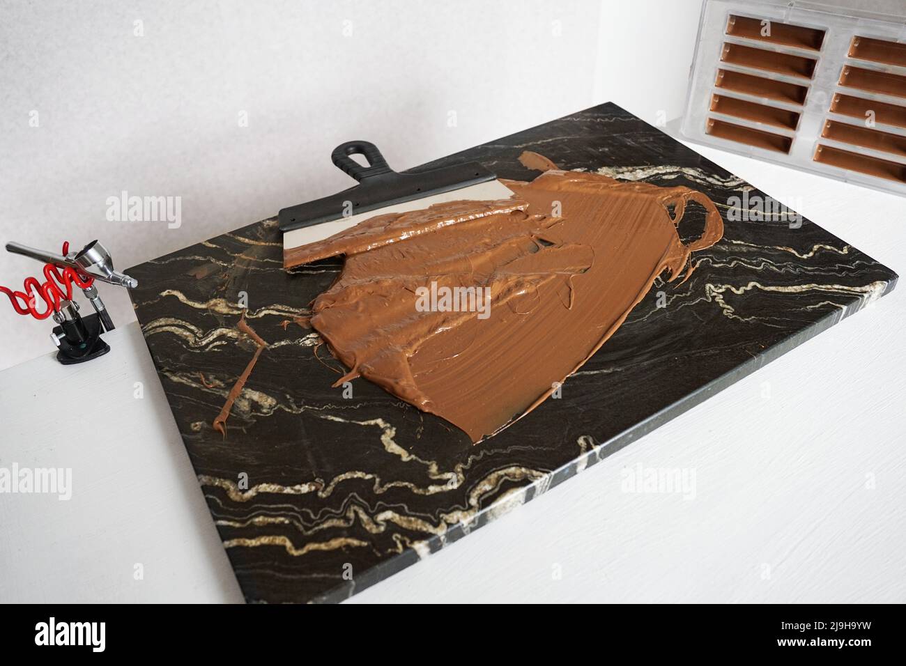 Milchschokolade auf einer Granitplatte zum Anlassen von Schokolade. Ein Spatel für die Verarbeitung von Schokolade. Eine Plastikform für Süßigkeiten auf dem Hintergrund. Stockfoto
