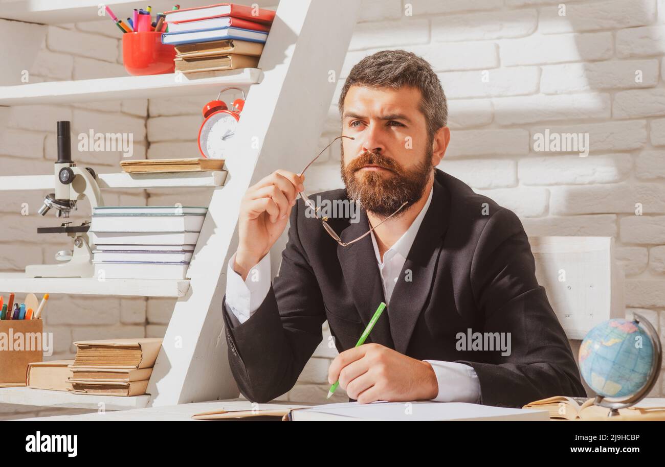 Porträt eines denkenden männlichen Lehrers im Klassenzimmer. Der Lehrer sitzt auf dem Schreibtisch und schreibt Text in einem Buch im Klassenzimmer. Stockfoto