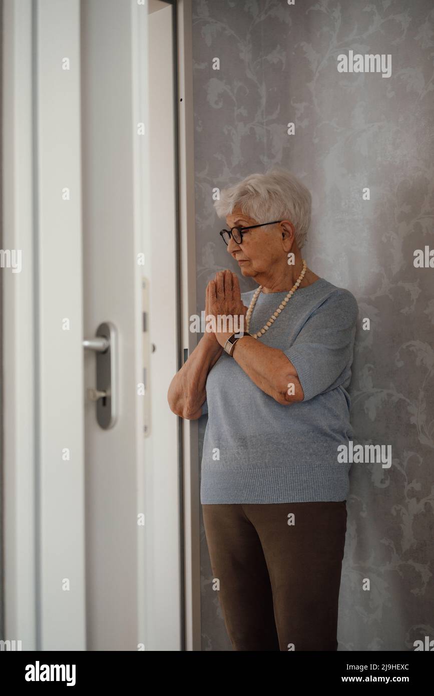 Ältere Frau mit zusammengekrallten Händen, die vor einer grauen Wand steht Stockfoto