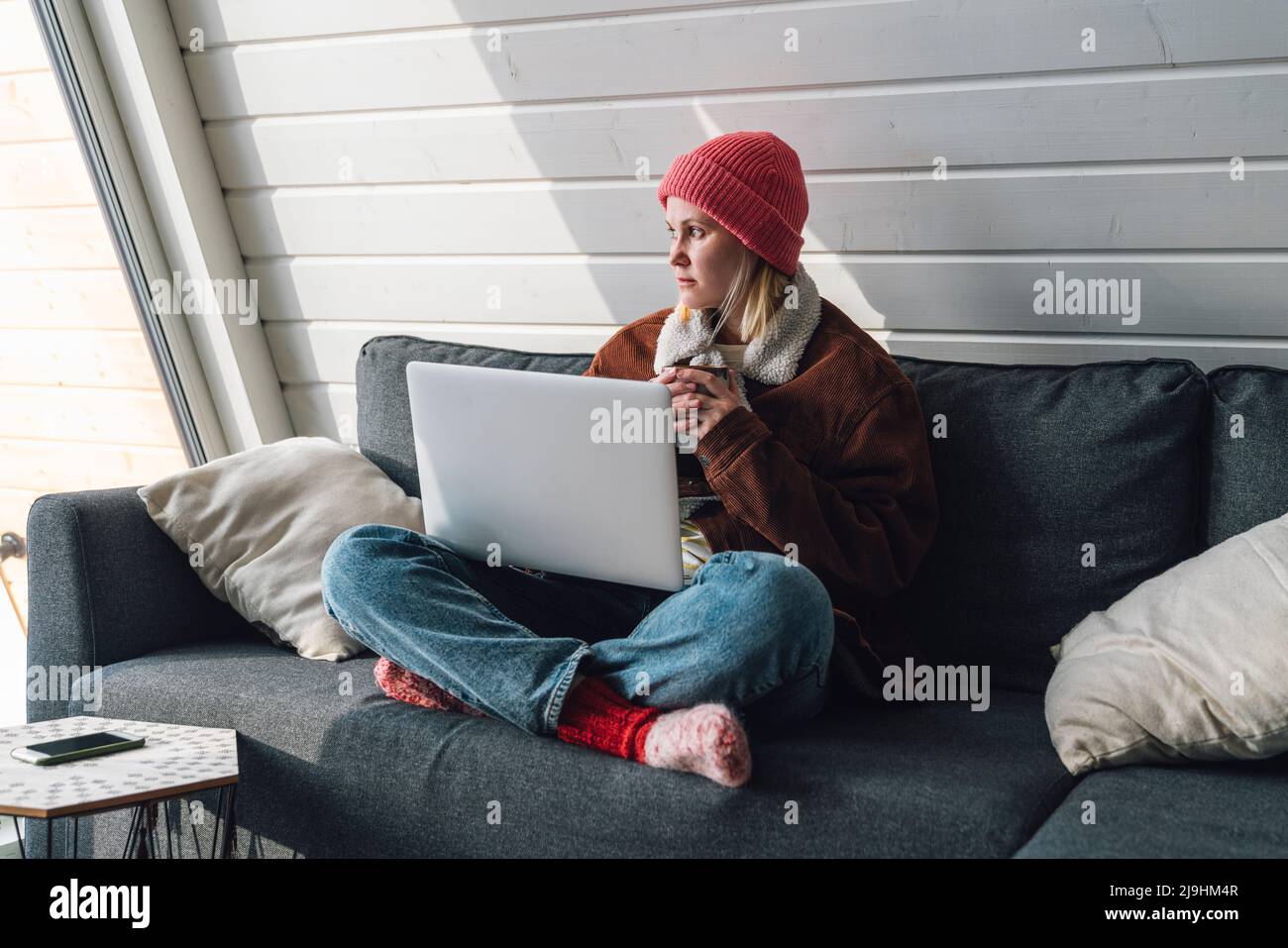 Frau in warmer Kleidung sitzt mit gekreuzten Beinen auf dem Sofa mit Laptop zu Hause Stockfoto