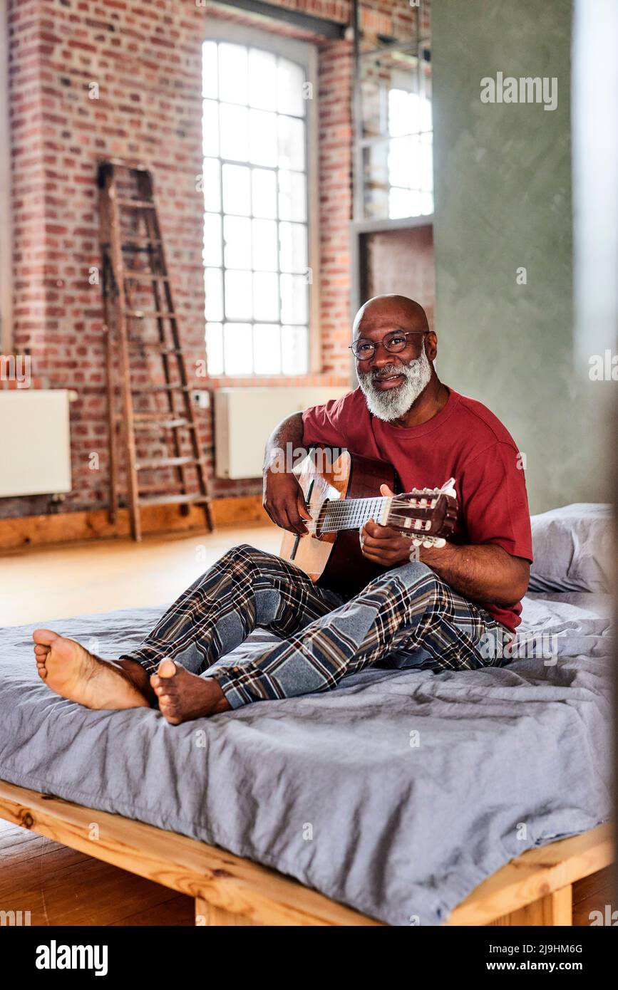 Reifer Mann mit Brille, der zu Hause Gitarre auf dem Bett spielt Stockfoto