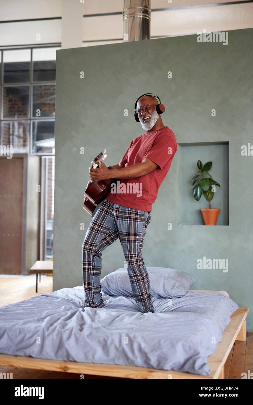 Reifer Mann mit Kopfhörern, der zuhause Gitarre auf dem Bett spielt Stockfoto