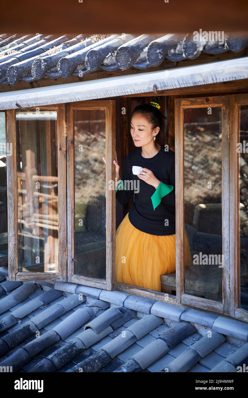 asiatische Frau, die Kaffee trinkt und aus einem traditionellen Haus aus dem Fenster blickt Stockfoto