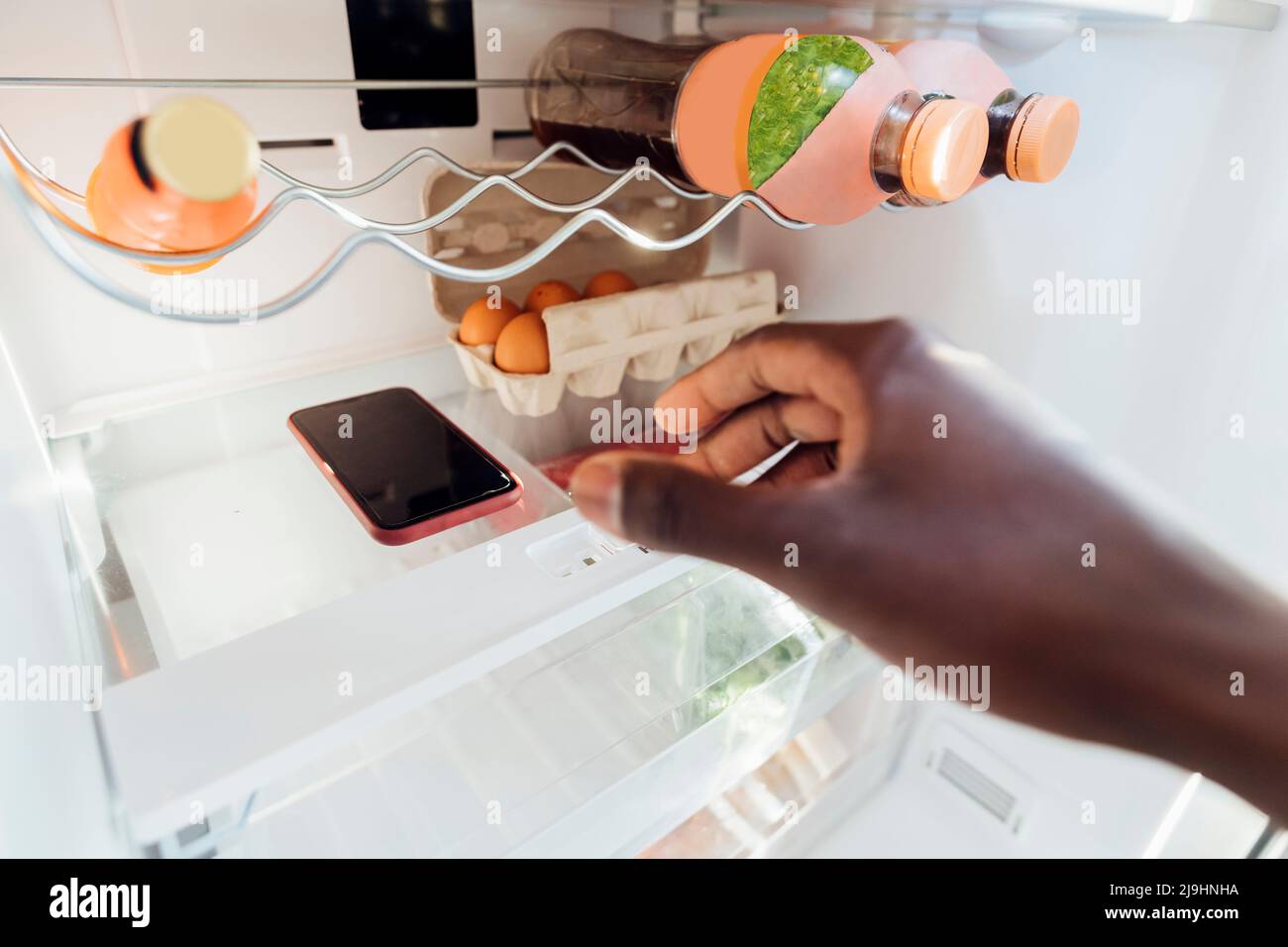 Hand des Mannes, der das Smartphone zu Hause im Kühlschrank hält Stockfoto