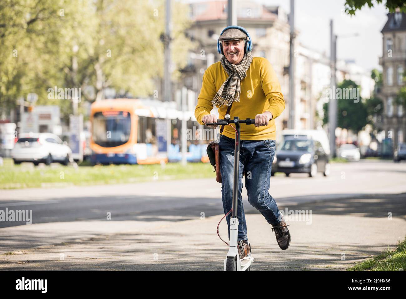 Glückliche ältere Männer mit kabellosen Kopfhörern fahren auf einem elektrischen Push-Roller Stockfoto