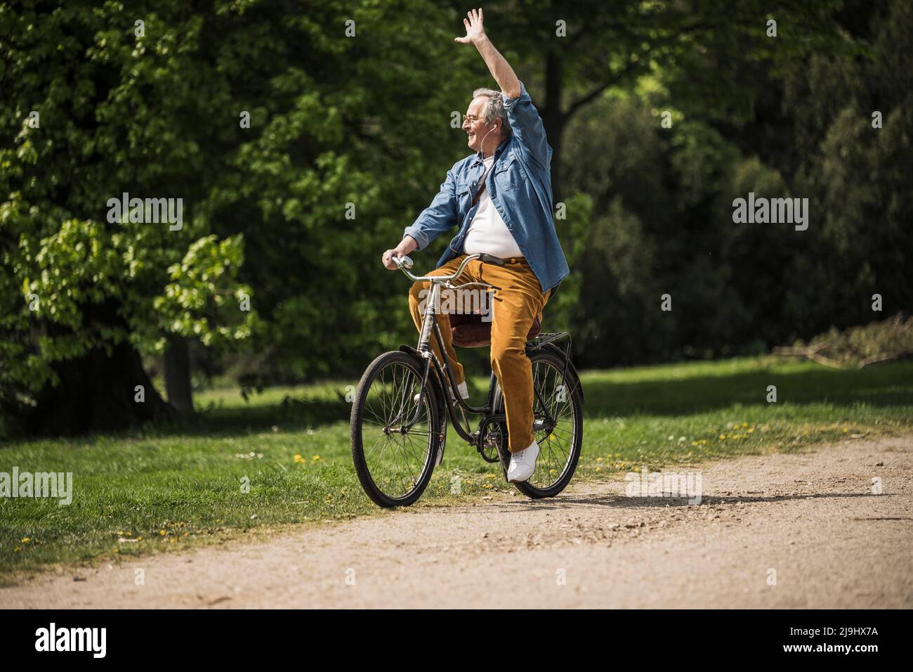 Lächelnder älterer Mann, der am sonnigen Tag mit dem Fahrrad im Park winkt Stockfoto
