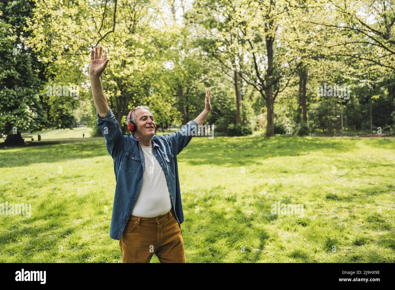Glücklicher älterer Mann mit erhobenen Armen und Musik über kabellose Kopfhörer im Park Stockfoto