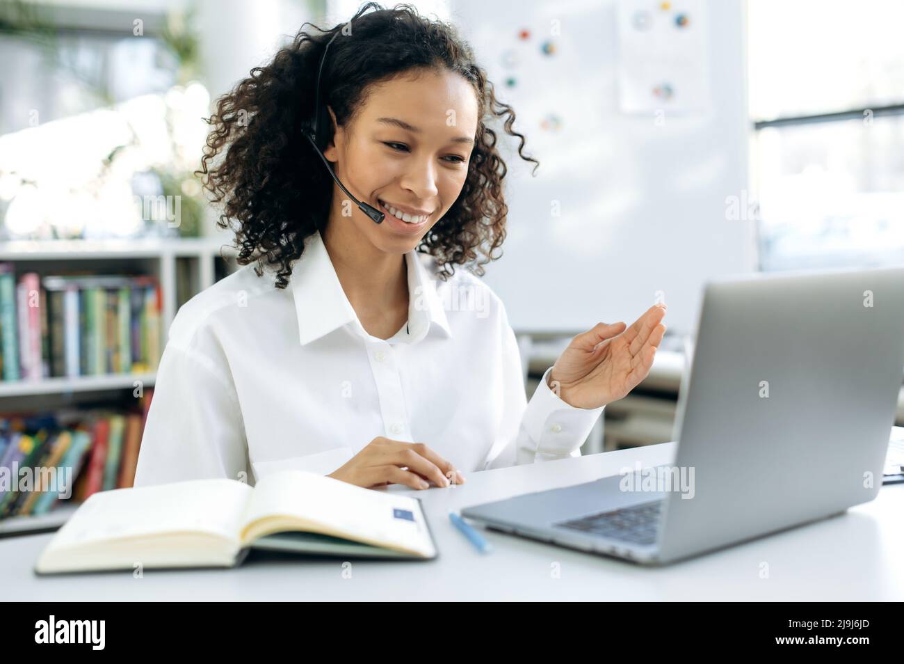 Erfolgreiche, smarte afroamerikanische Frau, Supportarbeiterin, Online-Consultant, Callcenter-Mitarbeiterin, sitzt an ihrem Schreibtisch vor einem Laptop, hat ein Video-Meeting mit einem Kunden, gestikuliert, lächelnd Stockfoto