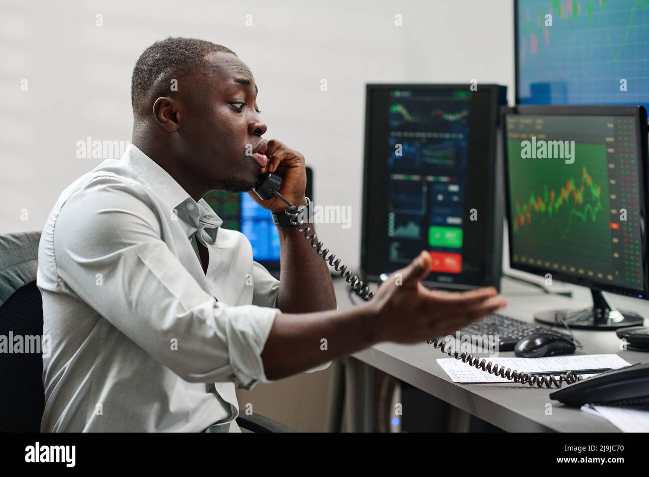 Emotionaler junger afroamerikanischer Mann, der in einer Makleragentur arbeitet und Probleme mit dem Kunden hat, der versucht, die Situation am Telefon zu erklären Stockfoto