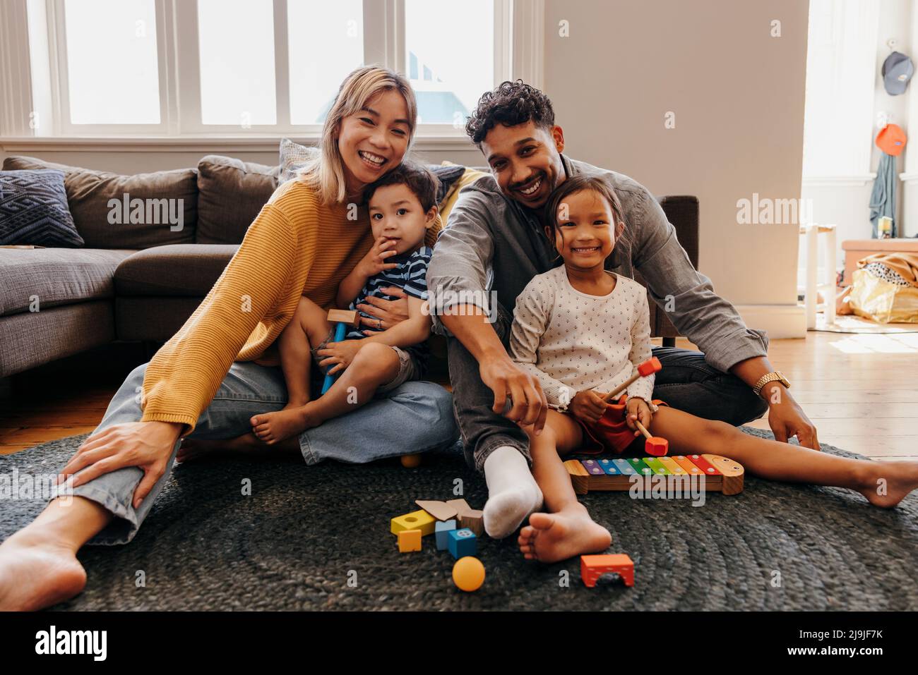 Junge Familie lächelt fröhlich in ihrem Wohnzimmer zu Hause. Zwei liebevolle Eltern, die während der Spielzeit mit ihrem Sohn und ihrer Tochter sitzen. Vierköpfige Familie Stockfoto