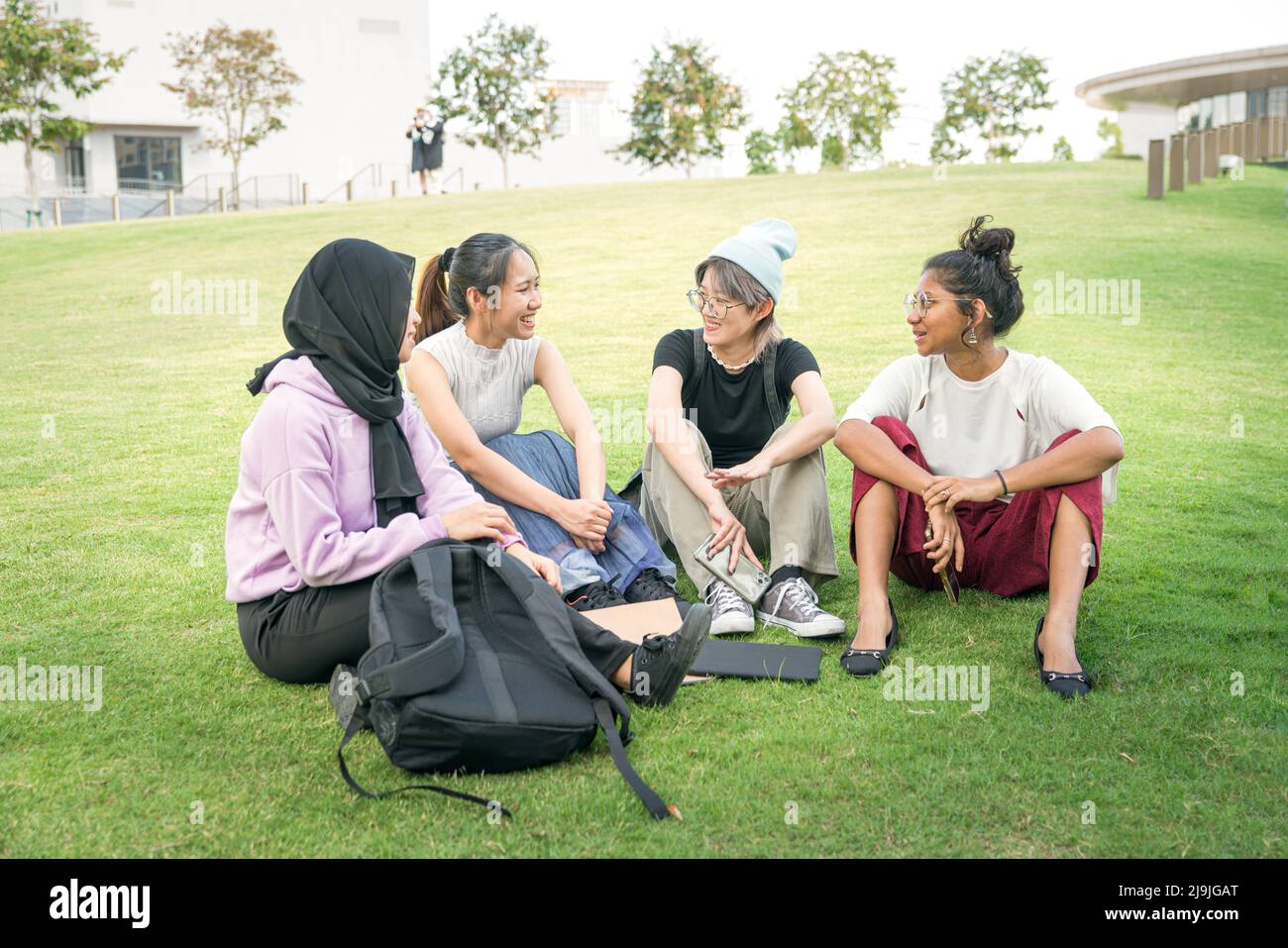 Eine Gruppe von vier jungen asiatischen Frauen, die auf grünem Rasen sitzen und sich unterhalten. College-Studenten mit Treffen und lockere Diskussion im Freien Konzept. Stockfoto