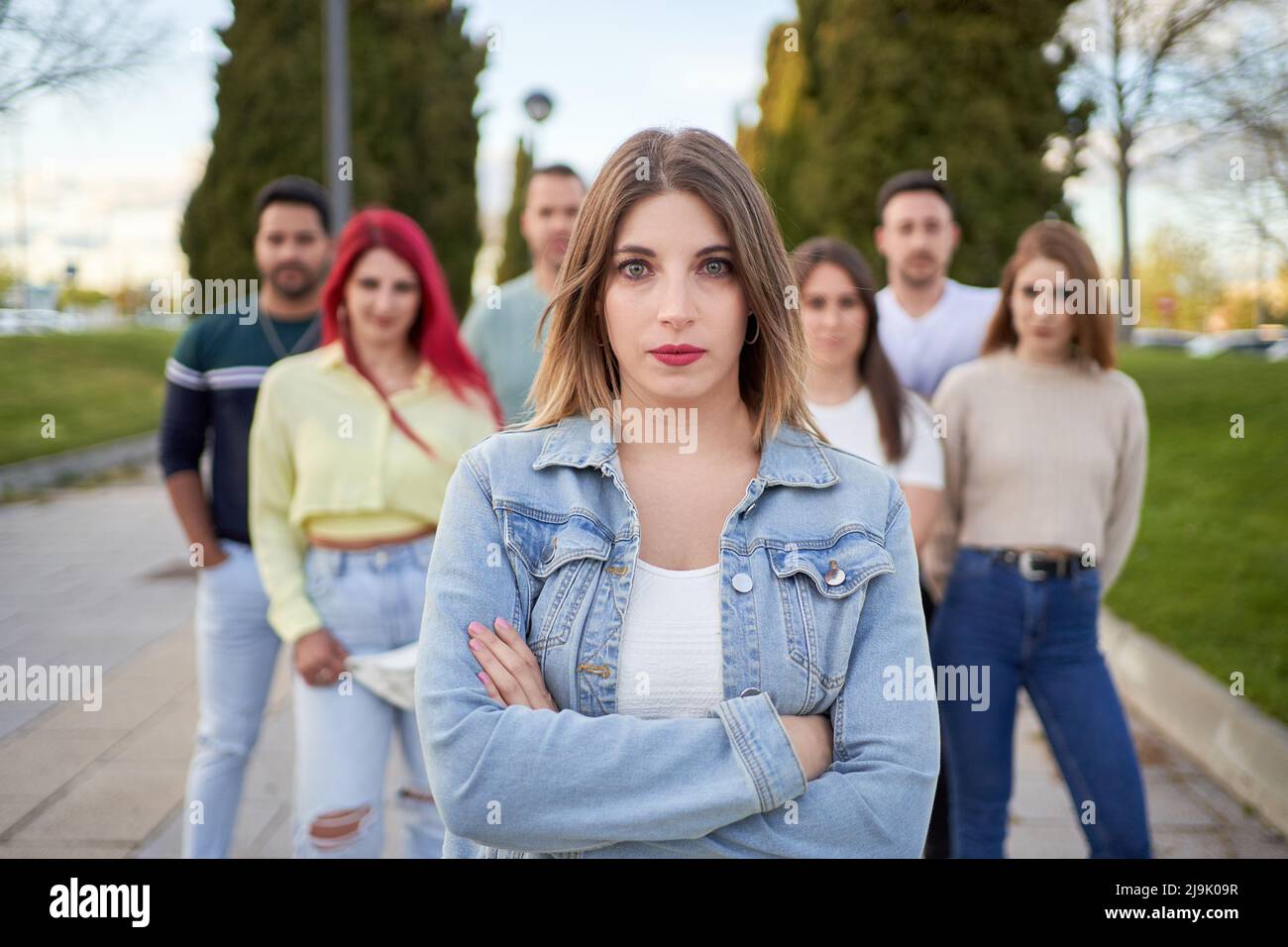 Selbstbewusste junge Frau mit gekreuzten Armen in Denim-Jacke, die auf die Kamera schaut, während sie auf der Straße gegen Freunde im Stadtpark steht Stockfoto
