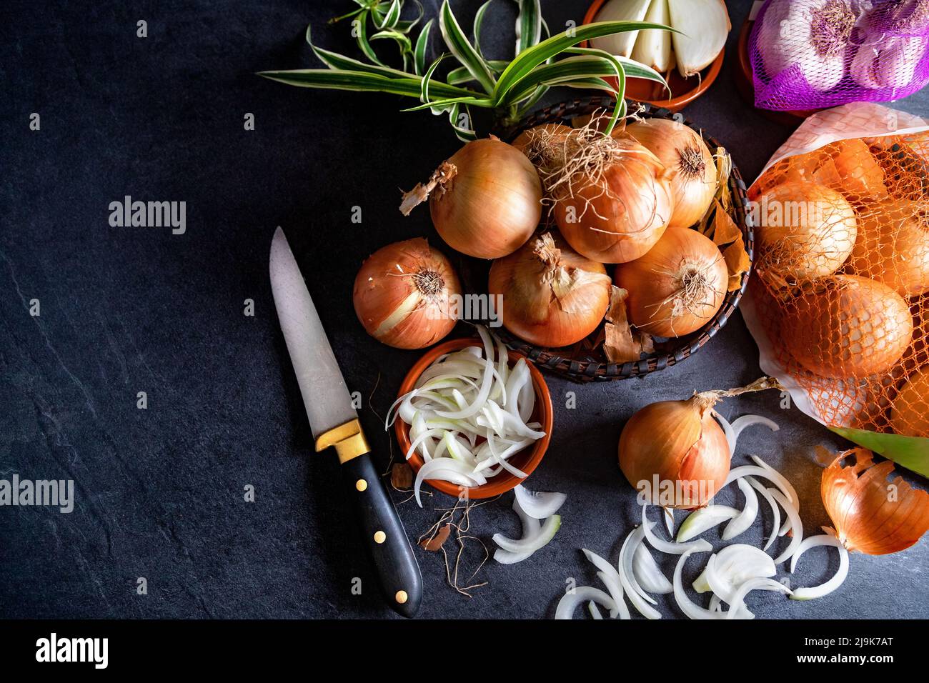 Eine Menge Zwiebeln in einem Korb und Teller auf einem dunklen und eleganten dunklen Strukturhintergrund. Zwiebeln und Scheiben auf Holzschneidebrett anbraten. Stockfoto