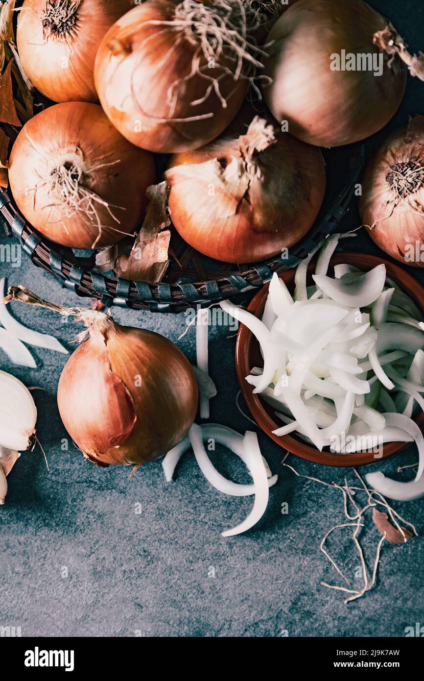 Eine Menge Zwiebeln in einem Korb und Teller auf einem dunklen und eleganten dunklen Strukturhintergrund. Zwiebeln und Scheiben auf Holzschneidebrett anbraten. Stockfoto