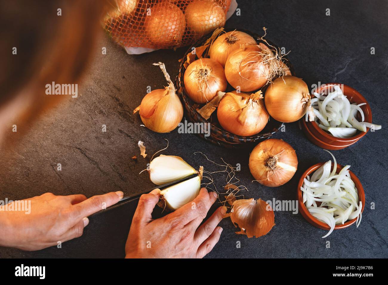 Viele Zwiebeln in einem Korb und Teller auf einem dunklen und eleganten dunklen Strukturhintergrund.Zwiebeln und Scheiben auf Holzschneidebrett anbraten. Stockfoto