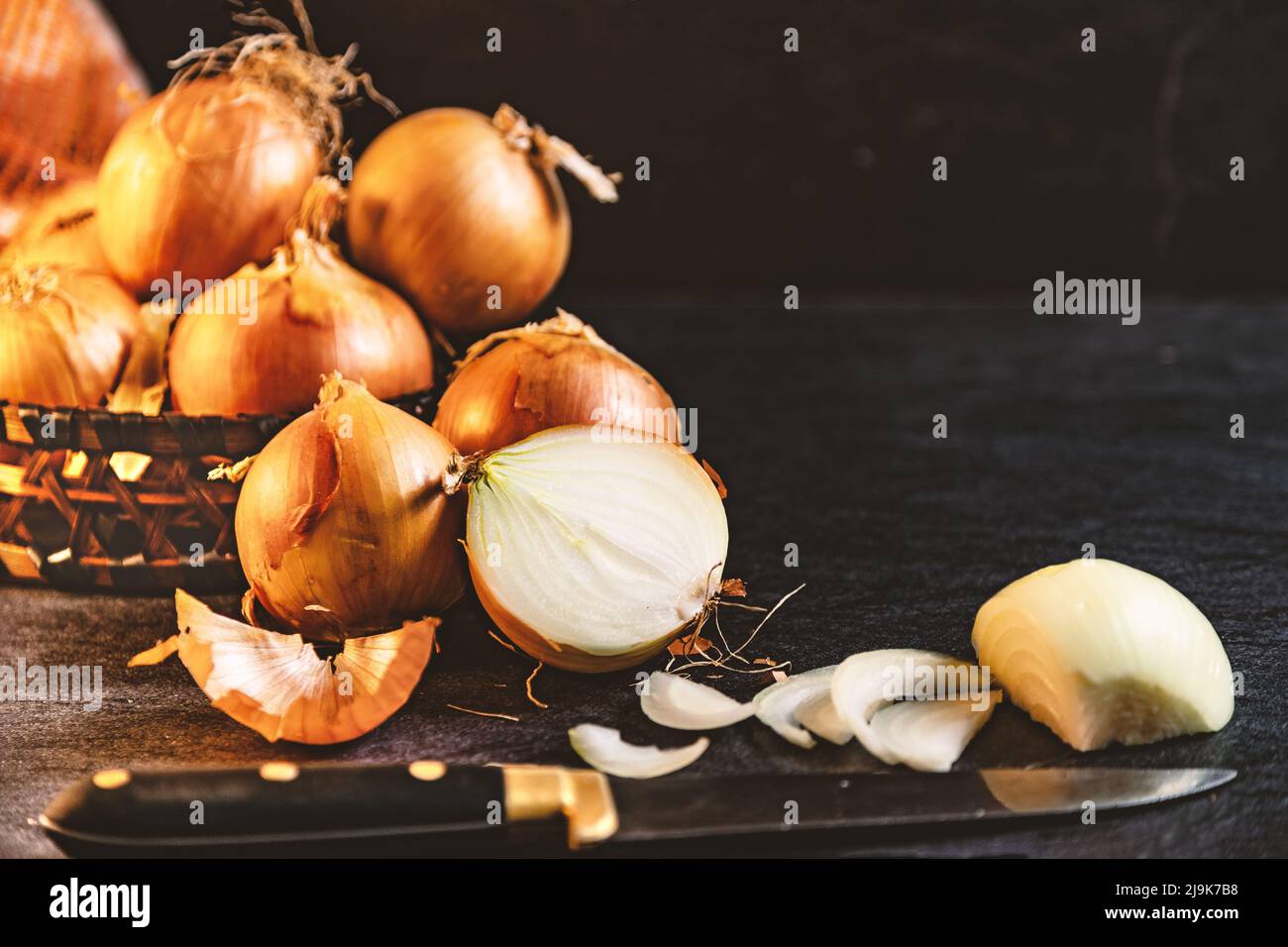 Eine Menge Zwiebeln in einem Korb und Teller auf einem dunklen und eleganten dunklen Strukturhintergrund. Zwiebeln und Scheiben auf Holzschneidebrett anbraten. Stockfoto