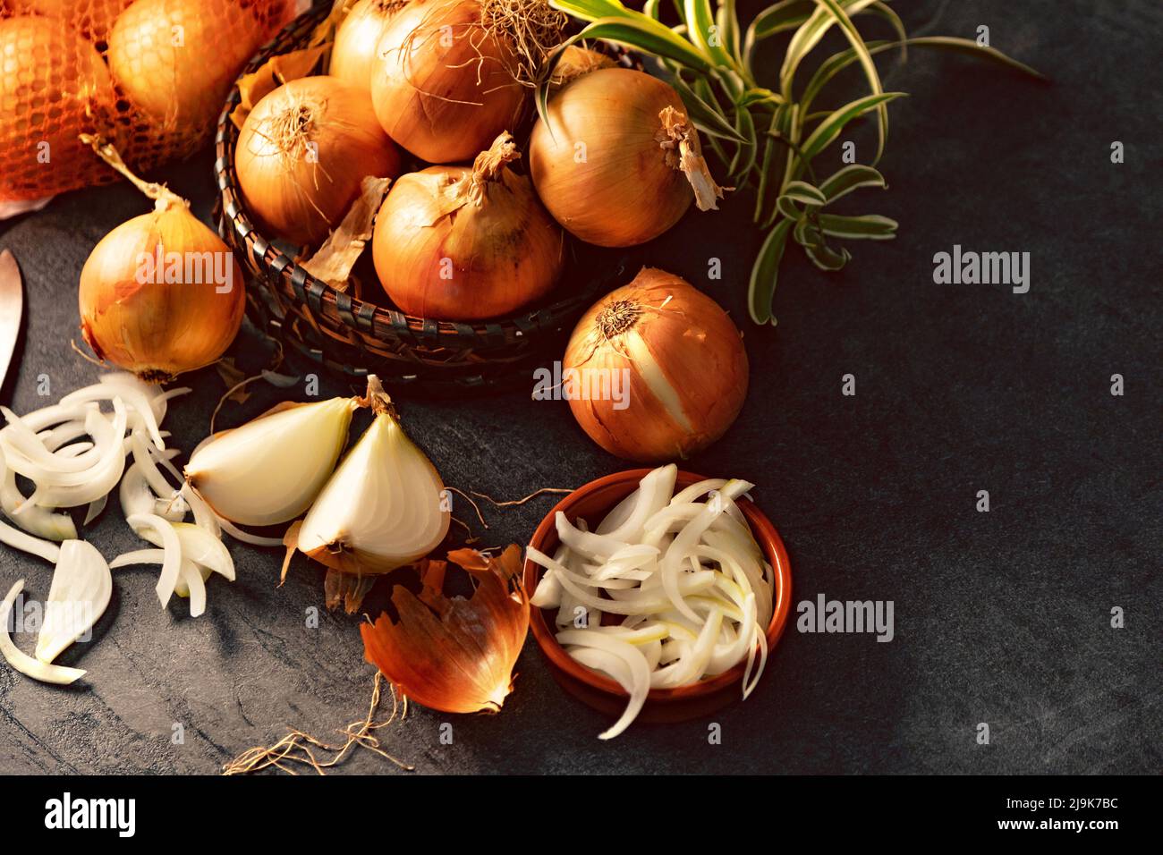 Eine Menge Zwiebeln in einem Korb und Teller auf einem dunklen und eleganten dunklen Strukturhintergrund. Zwiebeln und Scheiben auf Holzschneidebrett anbraten. Stockfoto