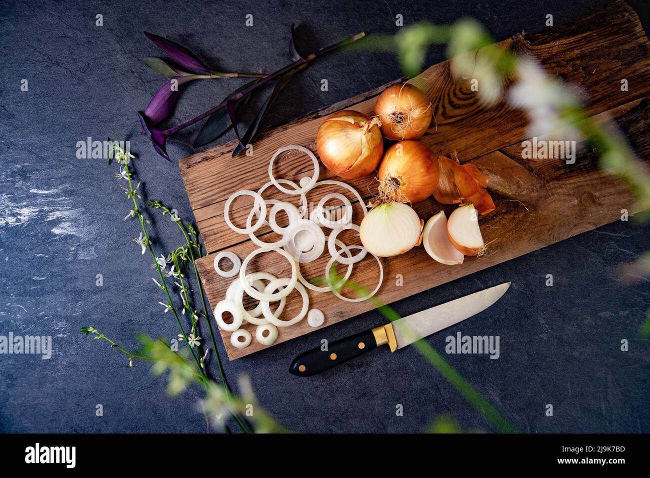 Eine Menge Zwiebeln in einem Korb und Teller auf einem dunklen und eleganten dunklen Strukturhintergrund. Zwiebeln und Scheiben auf Holzschneidebrett anbraten. Stockfoto