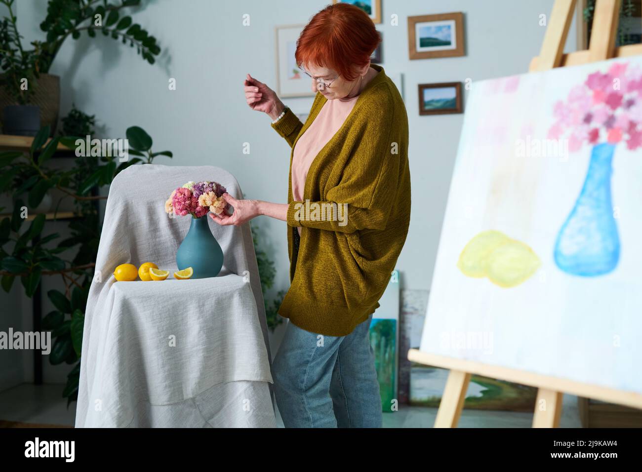Ältere Frau in legerer Kleidung korrigiert Blumen in der Vase, während sie Stillleben in der Klasse malt Stockfoto