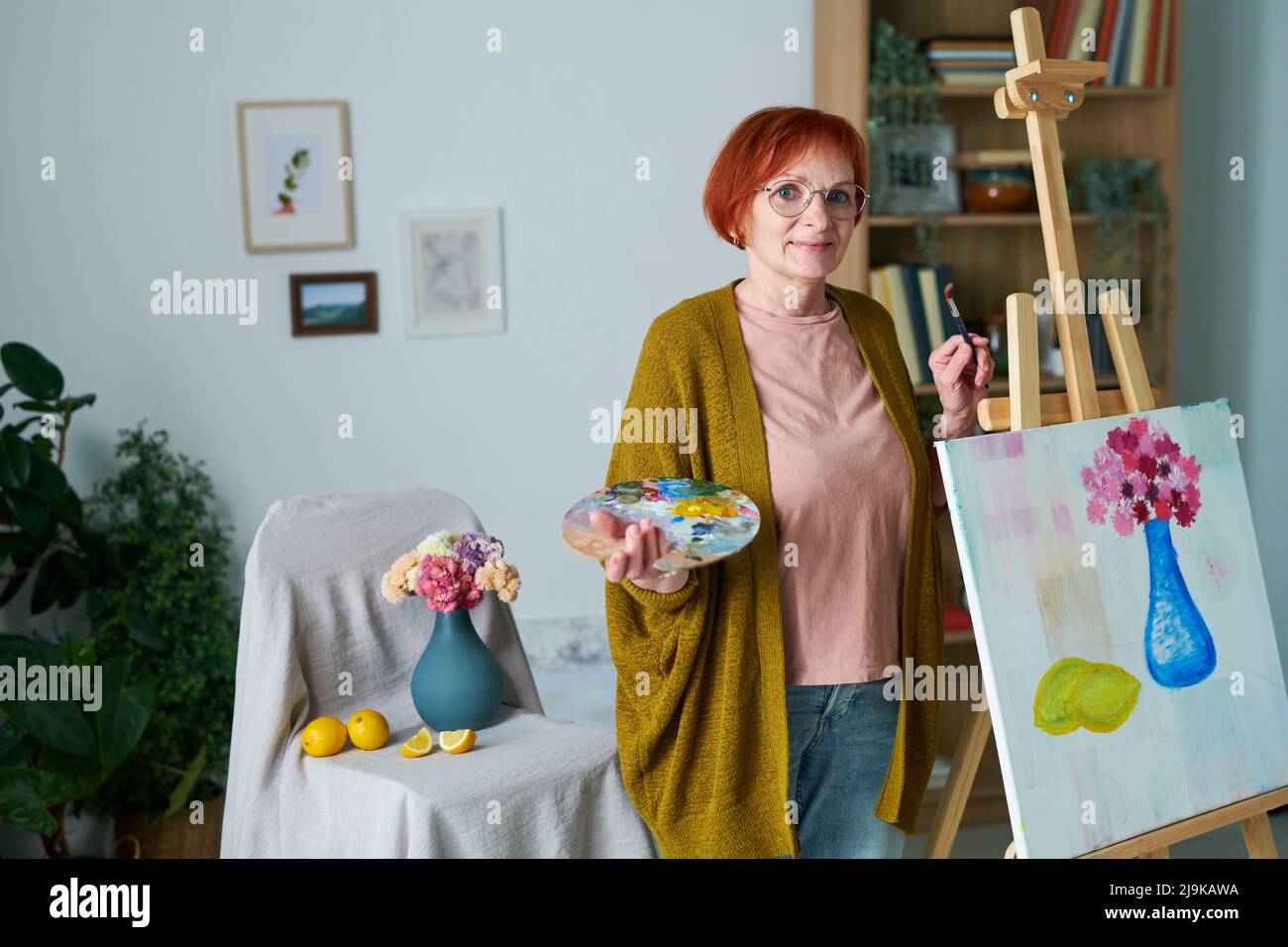 Porträt einer älteren Frau mit rothaarig in einer Brille, die die Kamera neben der Staffelei mit ihrem Bild im Zimmer betrat Stockfoto